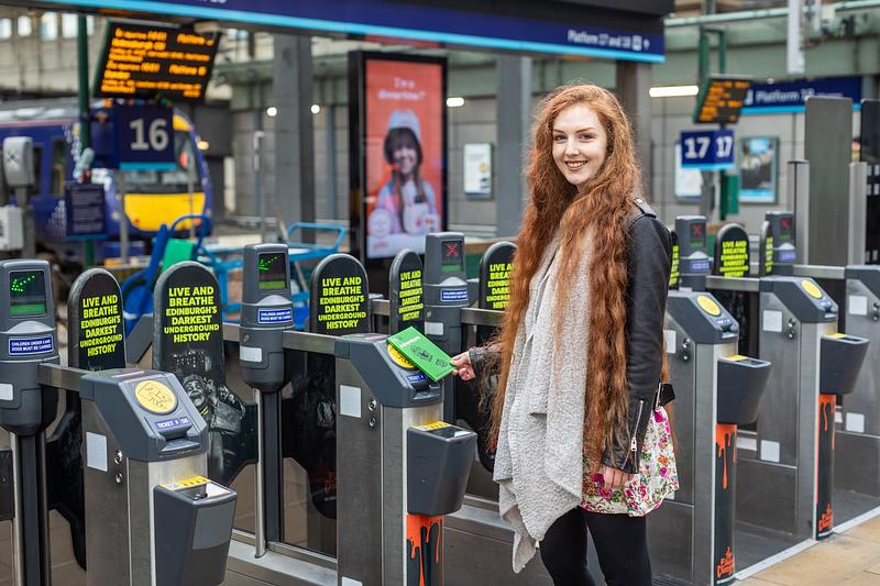 A young woman with waist length ginger hair stands at a set of train barriers